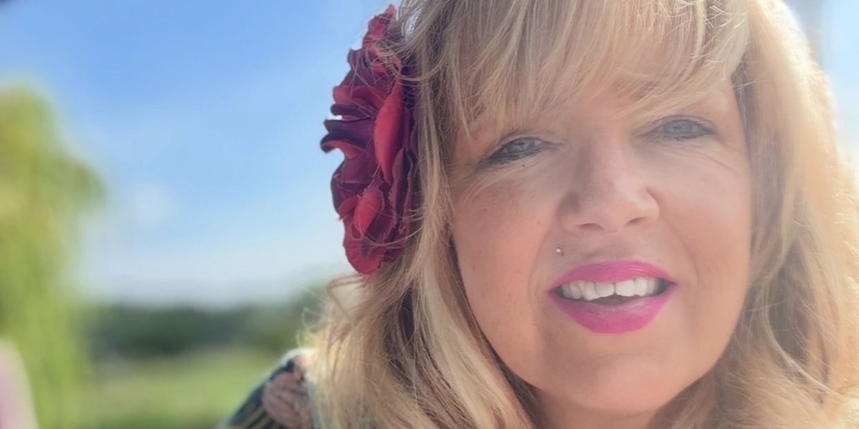 Smiling woman with blonde hair and a red flower in her hair outdoors.