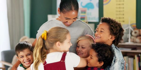A teacher shares a warm group hug with five joyful children in a colorful classroom.