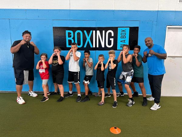 A group of kids and two coaches posing with boxing stances in front of a Boxing in a Box sign.