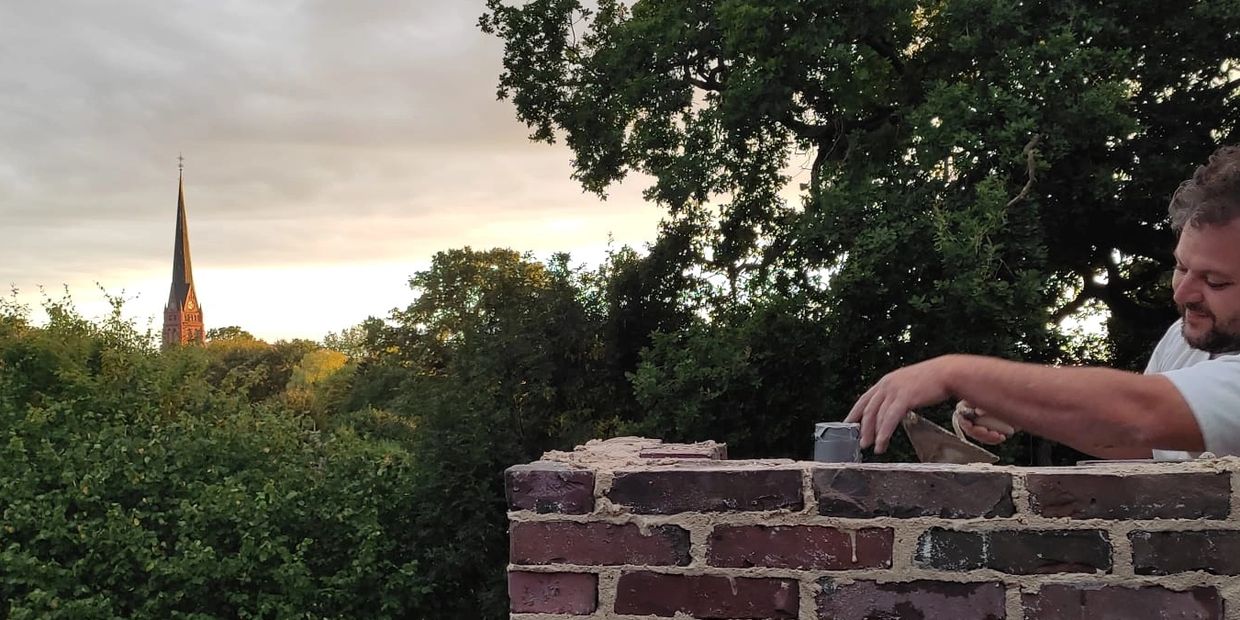 Mann mauert eine Ziegelmauer mit Bäumen und Kirchturm im Hintergrund.