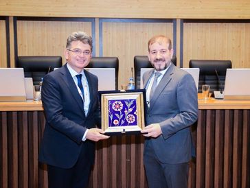 Two men in suits exchanging a framed flower artwork in a formal setting.