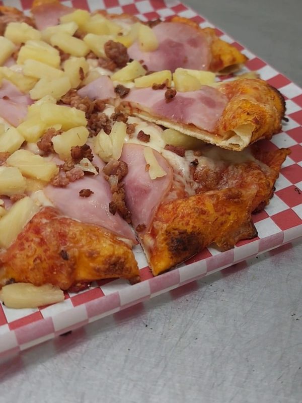 Close-up of a pizza with ham, pineapple, and ground meat on a checkered tray.