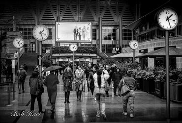 People walk under multiple clocks in a rainy urban setting with umbrellas.