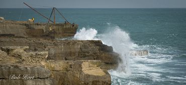 Waves crash against rocky cliffs by the sea under a cloudy sky.