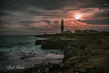 A lighthouse stands on a rocky coast at sunset under a cloudy sky.