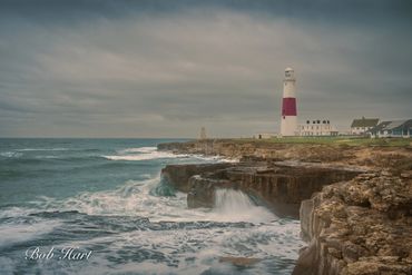 Lighthouse overlooking rocky cliffs and turbulent sea under cloudy sky.