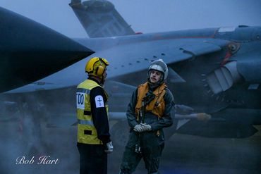 Two personnel stand near a military jet on a foggy day.