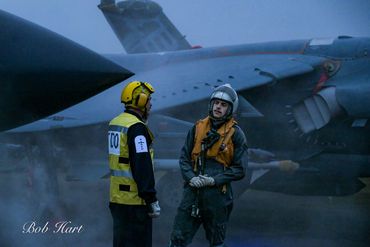 Two personnel stand near a military jet on a foggy day.