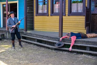 Cowboy aiming a rifle at a man lying down on porch steps.