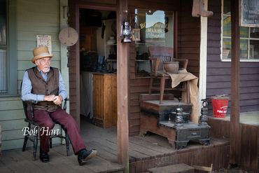 Old-fashioned man sitting on porch outside a rustic building.