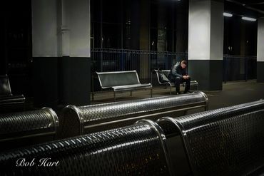 A solitary man waits on a dimly lit train platform at night.