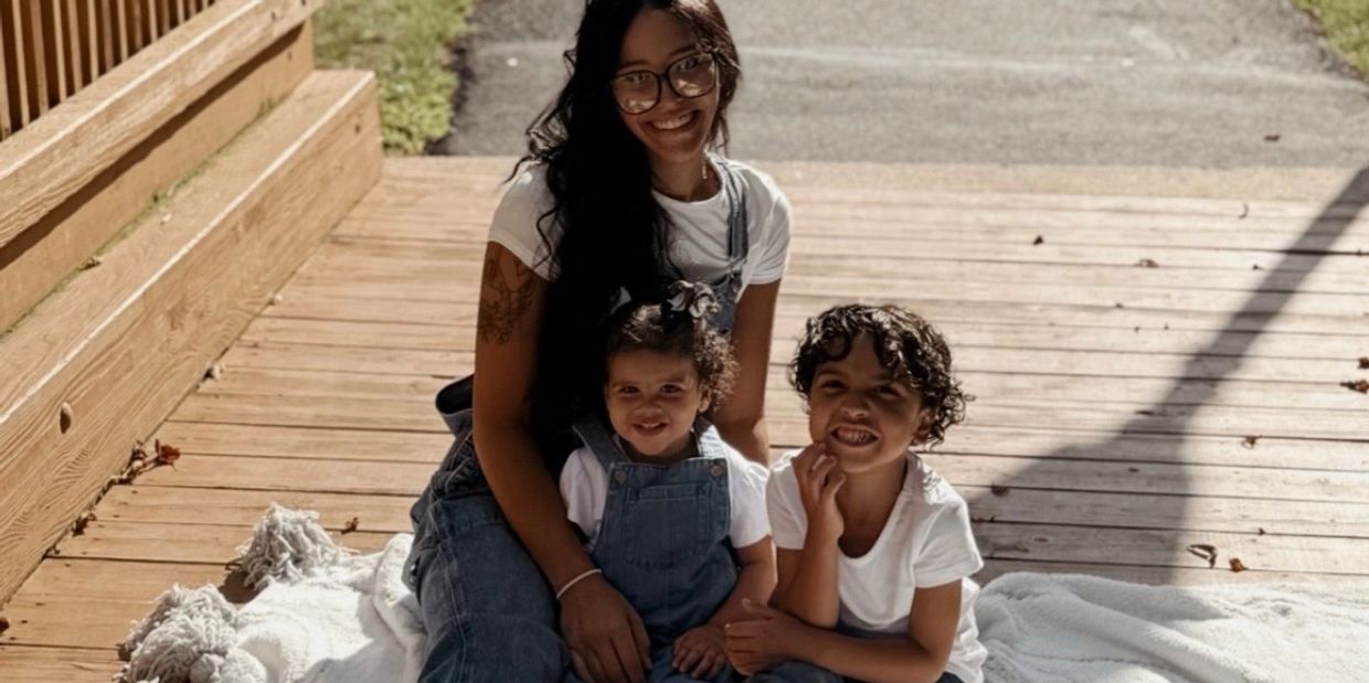 A smiling woman with two children sitting on a wooden deck with a white blanket.