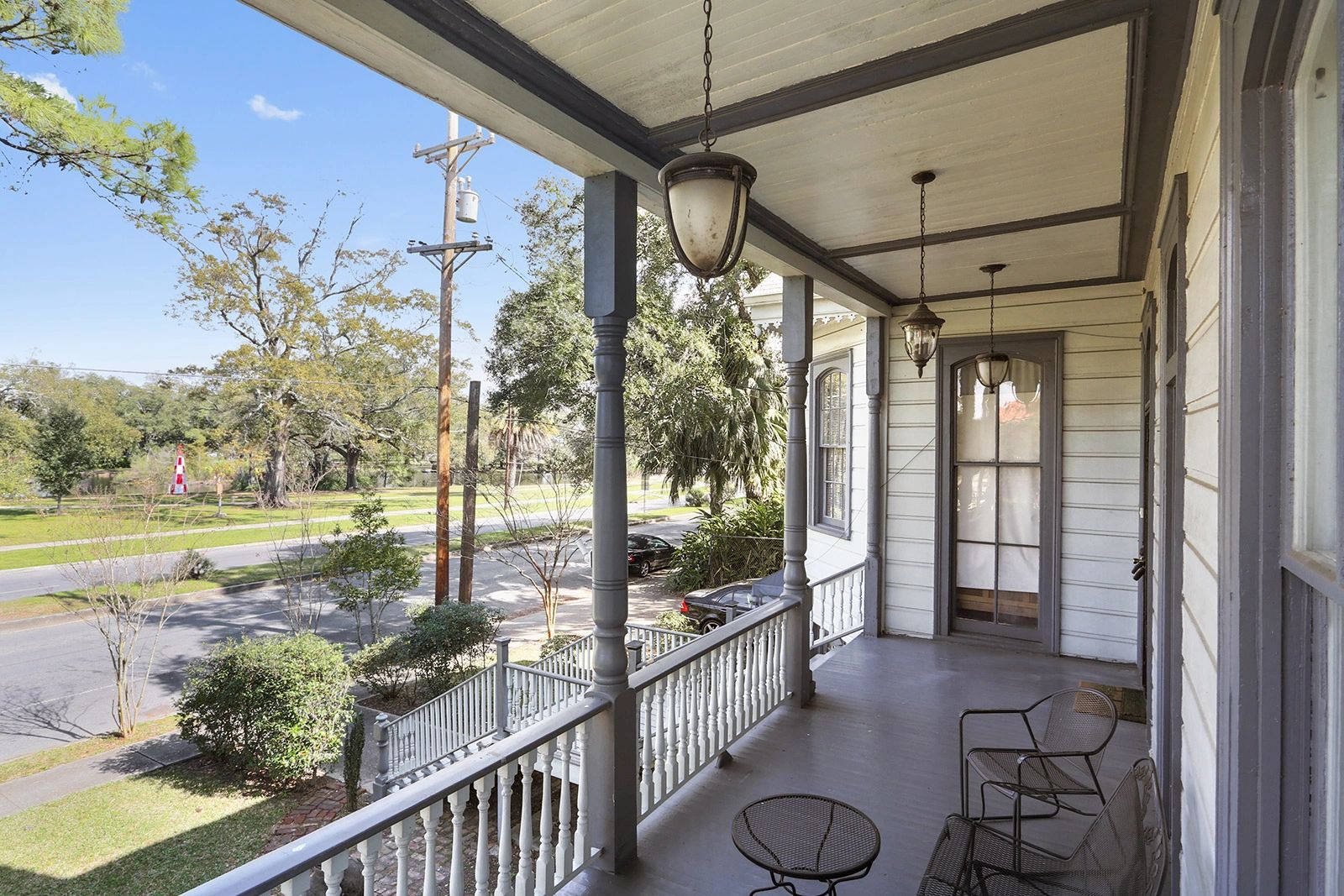 A covered porch with white siding and gray trim, overlooking a street with trees and a blue sky.