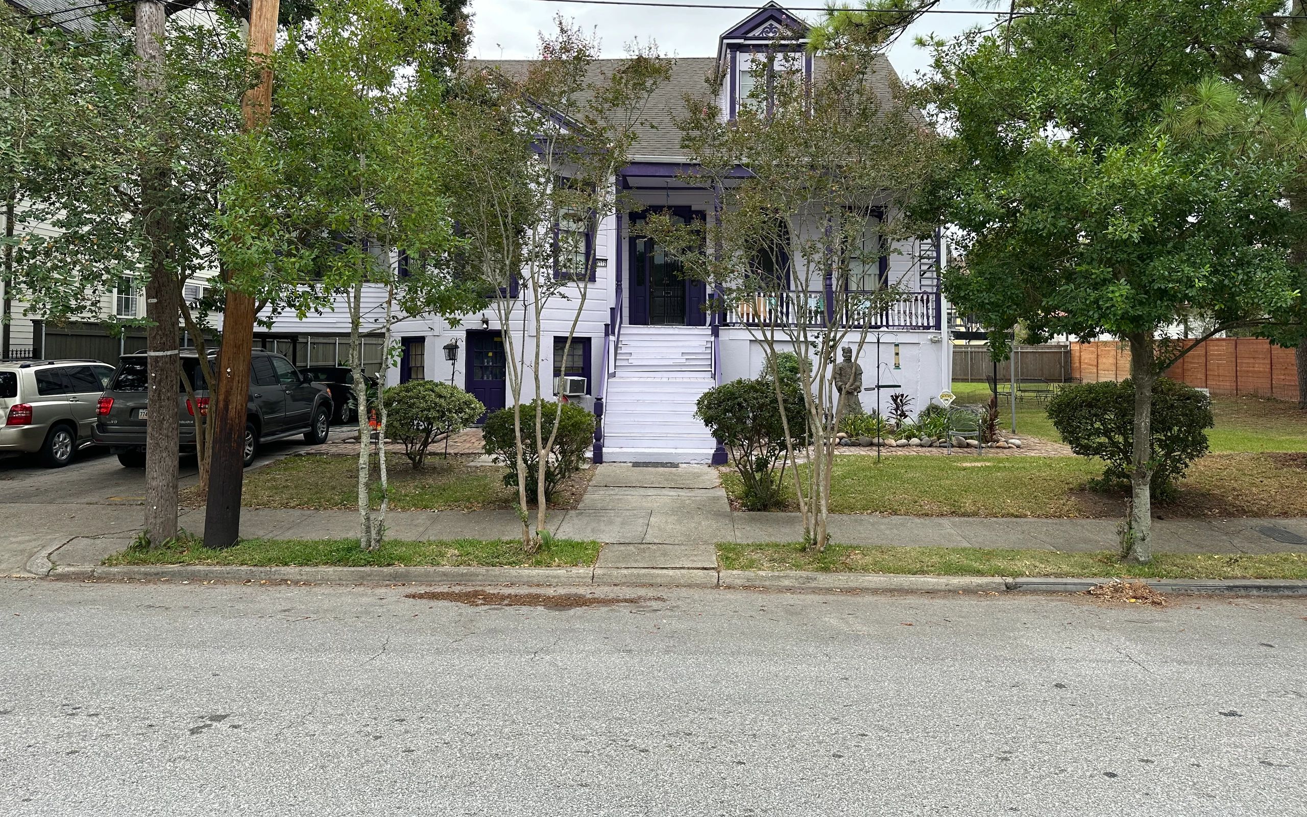 A white house with purple trim and a gray roof, surrounded by trees and a yard, with a sidewalk and road in front.