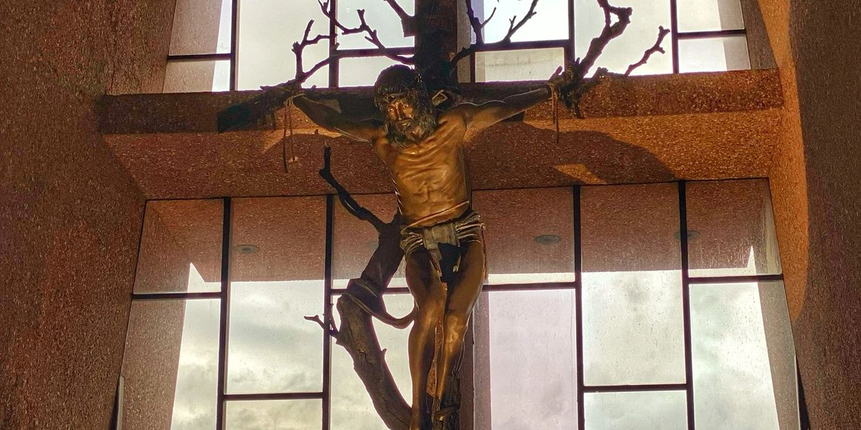 Bronze statue of Jesus on a tree-shaped cross inside a church window.