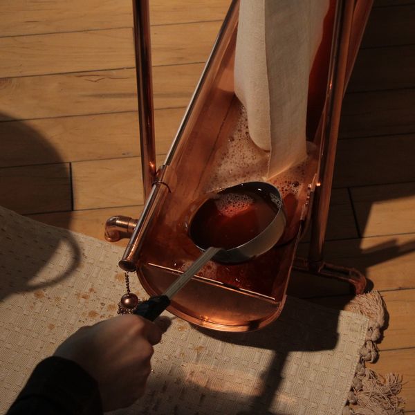 A hand holding a ladle over a copper container with liquid and a dripping cloth.