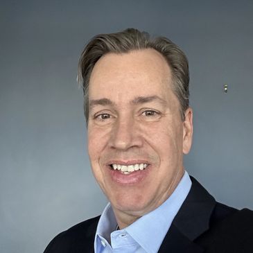 Smiling man in a blue shirt and black jacket against a plain gray background.