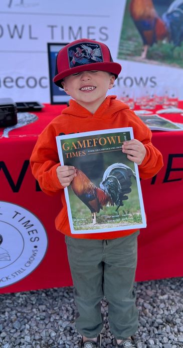 Smiling boy in red hoodie holding a Gamefowl Times magazine at an outdoor event.