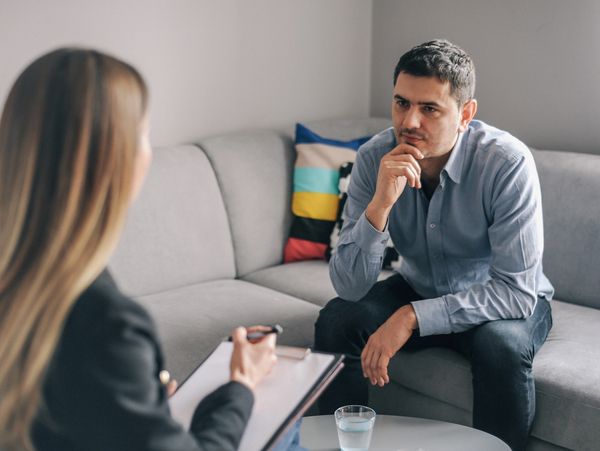 A man with a pensive expression sits on a sofa, looking at a woman who is taking notes.