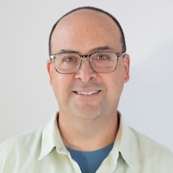 Smiling man wearing glasses and a light-colored shirt against a plain background.