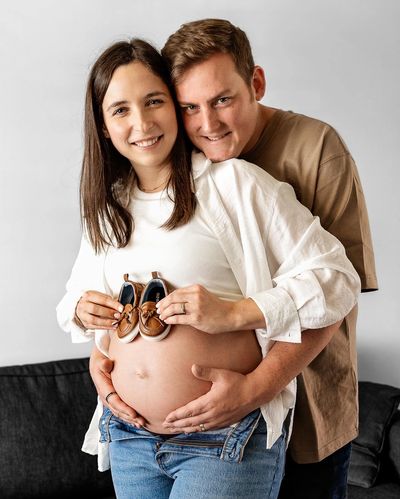 Pregrant lady holding baby shoes up to her belly while husband wraps his arms around her from behind
