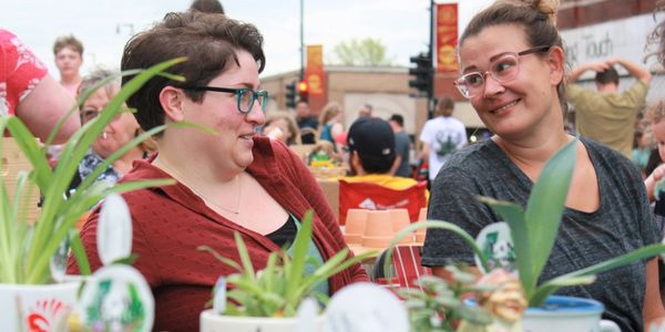 Two women smiling and chatting at an outdoor market with plants in the foreground.