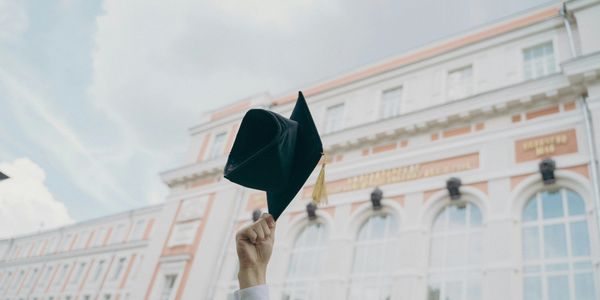 Student holding up a graduation cap in front of a historic building.