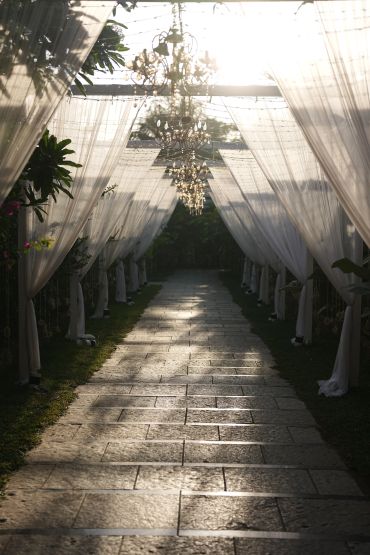 Sunlit pathway with hanging chandeliers under sheer curtains.