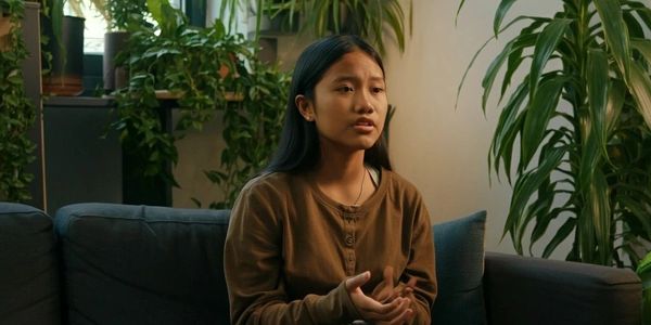 Young woman sitting on a couch surrounded by indoor plants, speaking thoughtfully.