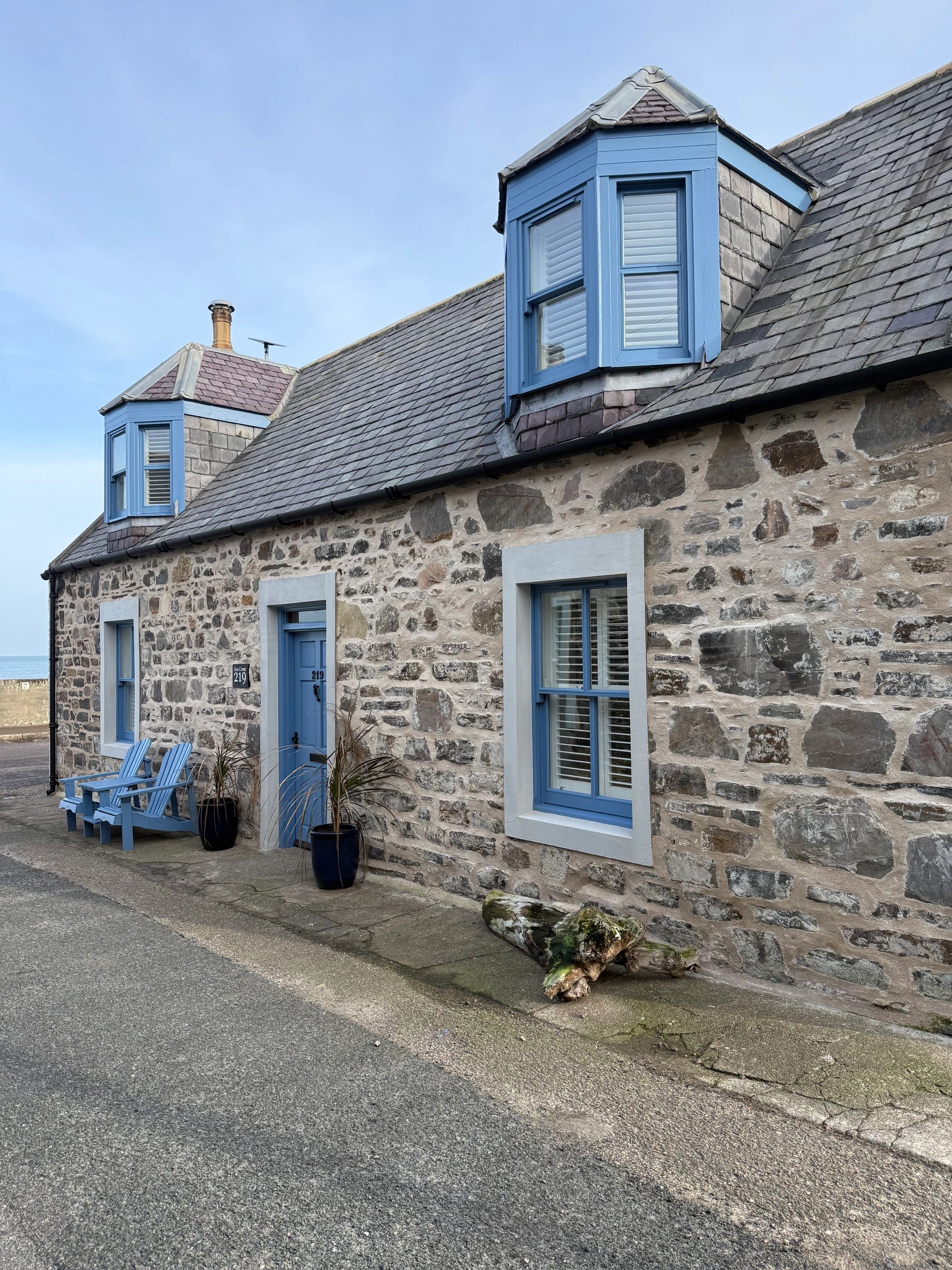 Stone cottage with blue windows and chairs by the road.