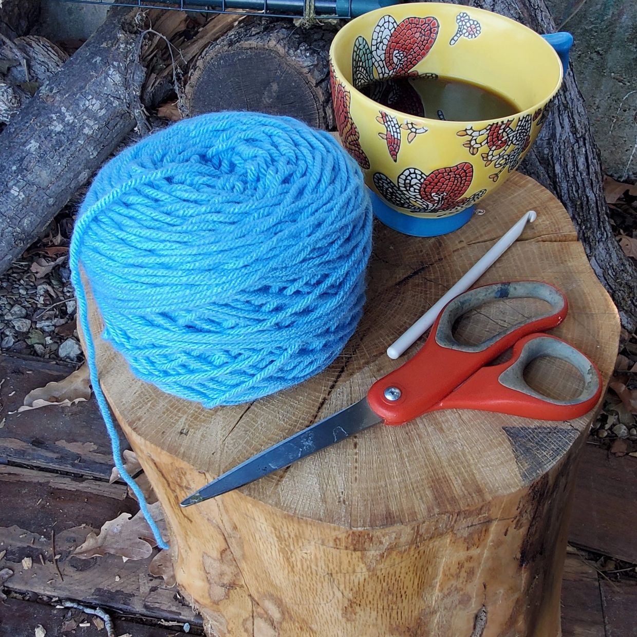A cake of bright blue yarn, a pair of red scissors, a crochet hook, and a cup of coffee on a stump
