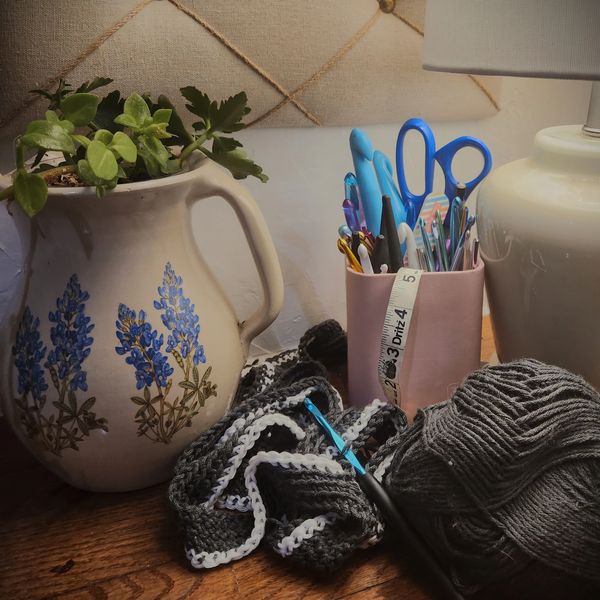 A craft table with ceramic pot, scissors, crochet hooks, measuring tape, yarn, and crochet project .
