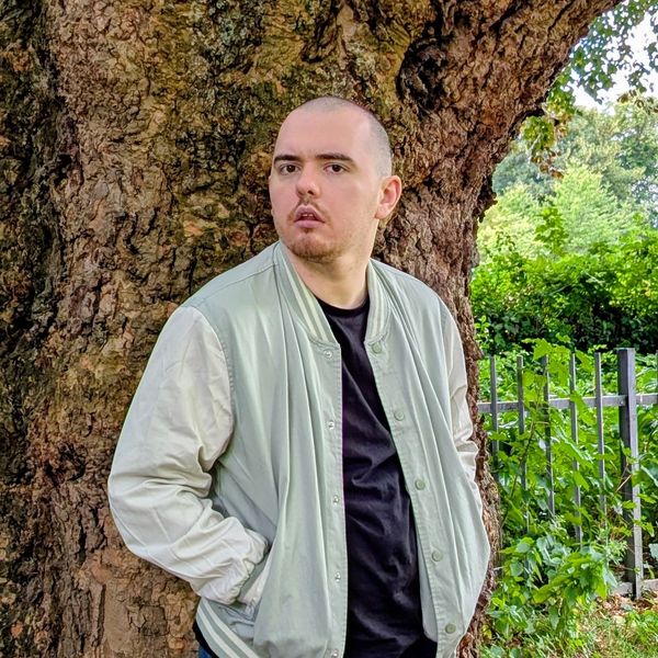 Man in casual jacket standing by a large tree in a park.