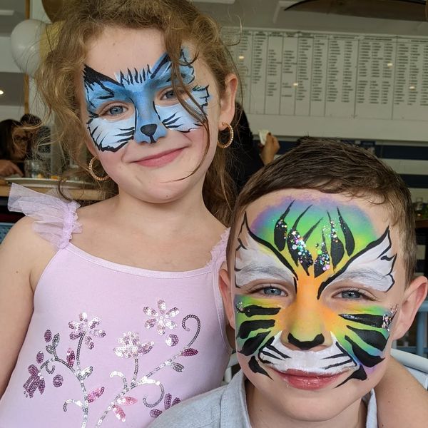 Two children with colorful tiger face paint smiling together indoors.