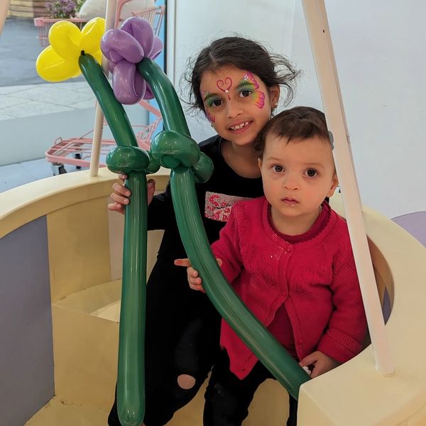 Two children with face paint holding balloon flowers, one smiling and one curious.