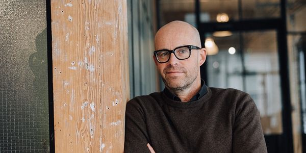 Bald man with glasses leaning against a wooden pillar indoors.