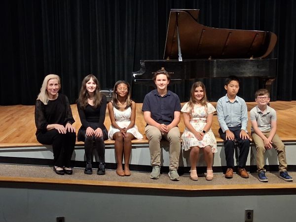 Group of children and an adult posing on stage with a grand piano.