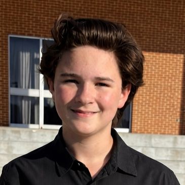 Smiling young person with short brown hair in front of a brick building.