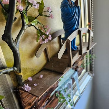 A miniature wooden bridge with pink flowers and a blue statue in the background.