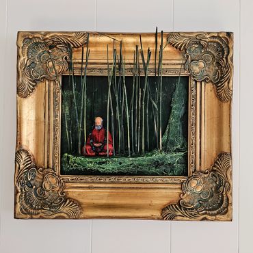 Miniature figure meditating among bamboo in ornate gold frame.