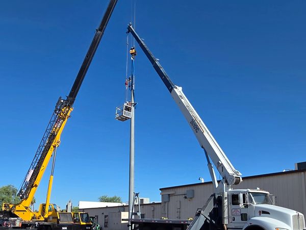 Two cranes lifting a pole with a basket under a clear blue sky.