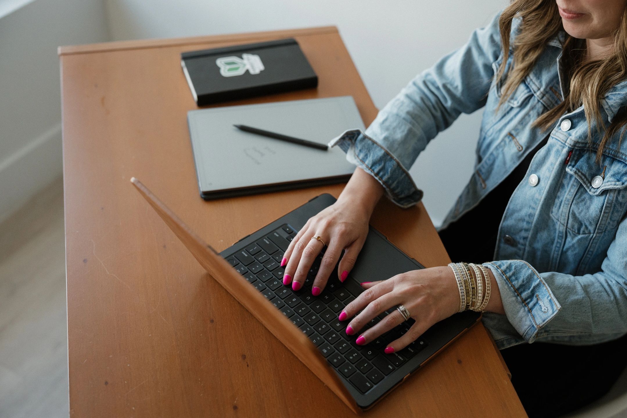 Woman typing on a laptop at a wooden desk wearing a denim jacket.