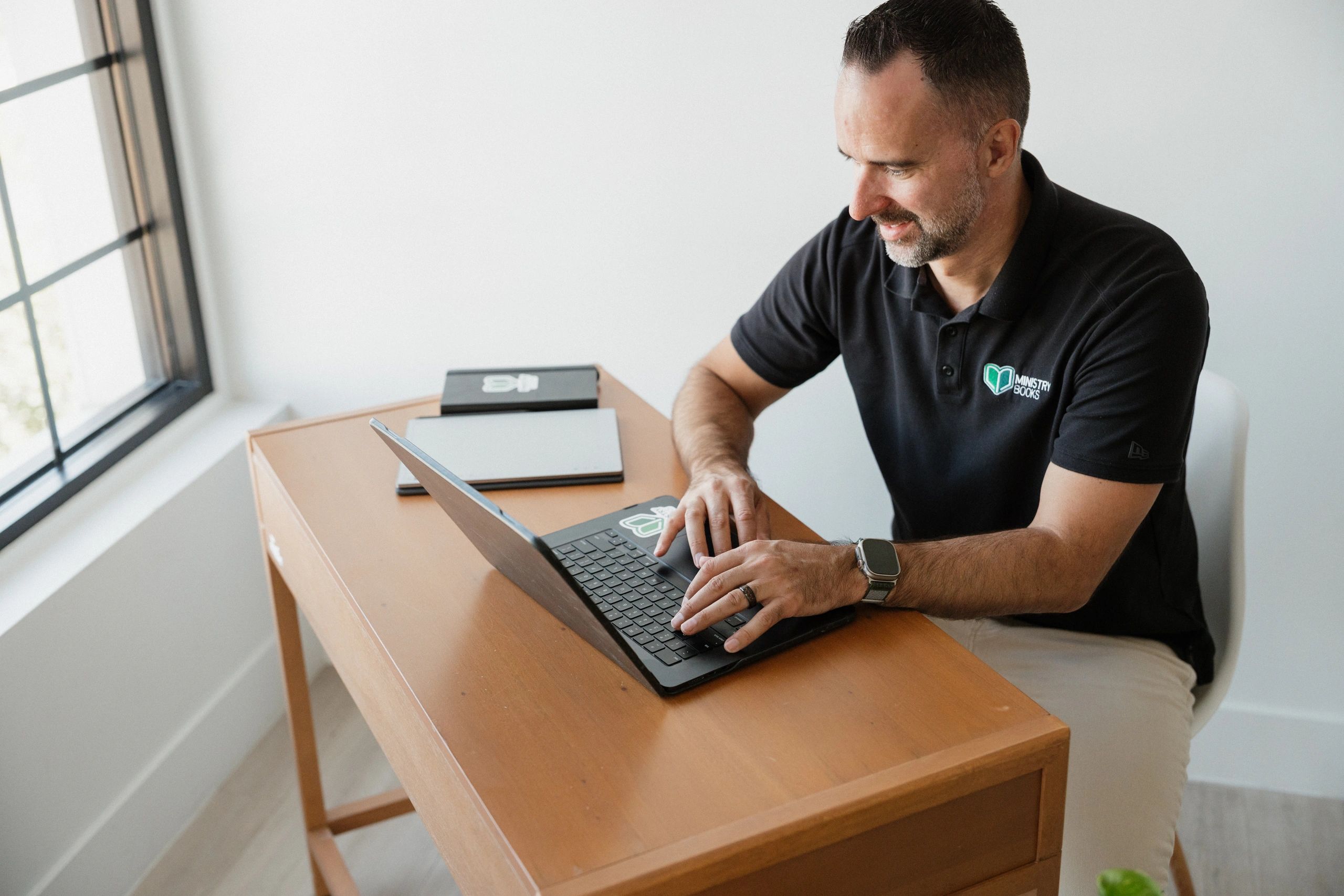 Man working on laptop at wooden desk near window.