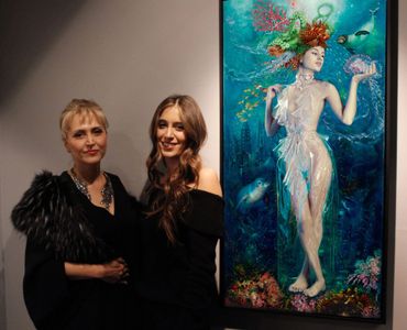 Two women posing beside a vibrant underwater-themed painting at an art gallery.