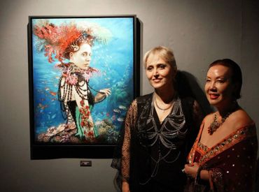 Two women posing next to a colorful underwater-themed portrait in an art gallery.