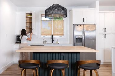 Modern kitchen with wooden island and woman reaching for a cup in cabinet.