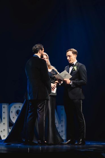Two men and one woman in formal attire on stage, reading from papers.
