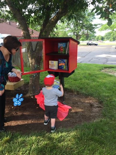 A child picks a book from a little free library while an adult watches nearby.