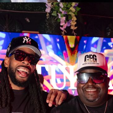 Two men smiling, wearing sunglasses and hats indoors with colorful background.