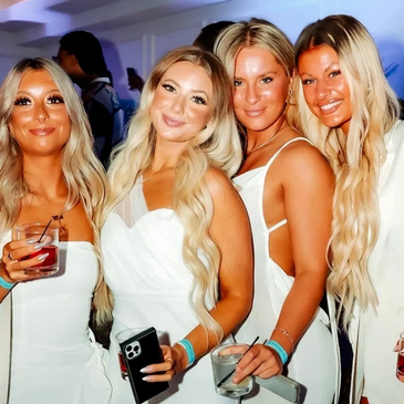 Four women in white dresses smiling and holding drinks at a party.